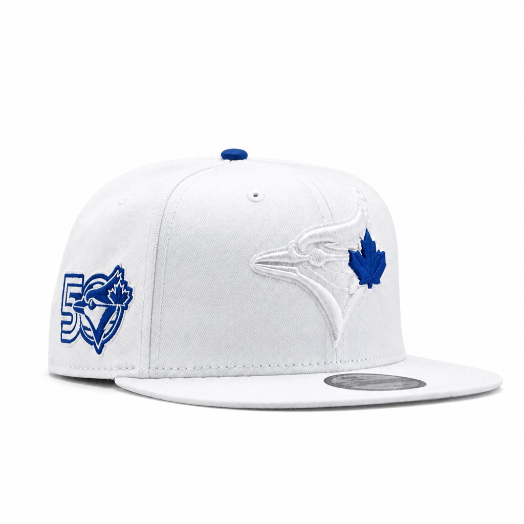 A white structured baseball cap with blue embroidered details, featuring the Toronto Blue Jays logo on the front and side, and a blue button on top, photographed on a clean white background.
